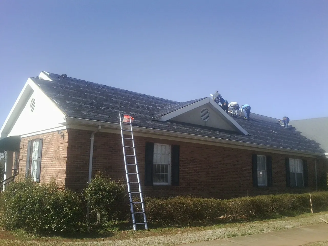 Crew tearing shingles off a brick ranch roof with a ladder set — Metal Roof Repair work by our crew in East Otis