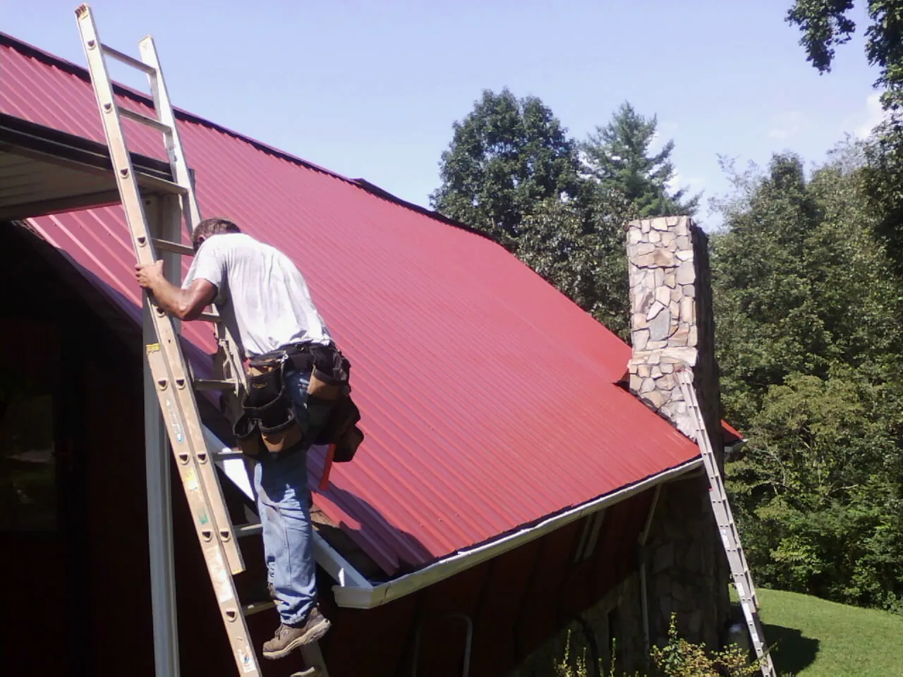 Roofer on a ladder beside a red metal roof and stone chimney — Soffit & Fascia Repair by Roofing Brothers in East Otis